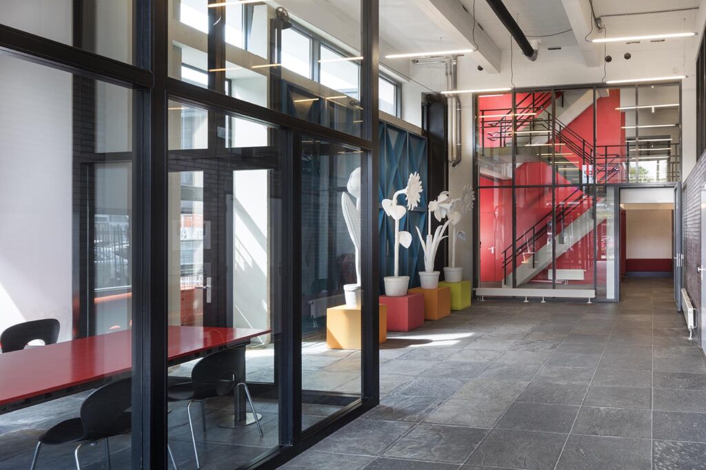 Moderne entreehal aan de Generaal Vetterstraat met glazen wanden, kleurrijke accenten, opvallende trappen en kunstzinnige bloemeninstallatie.